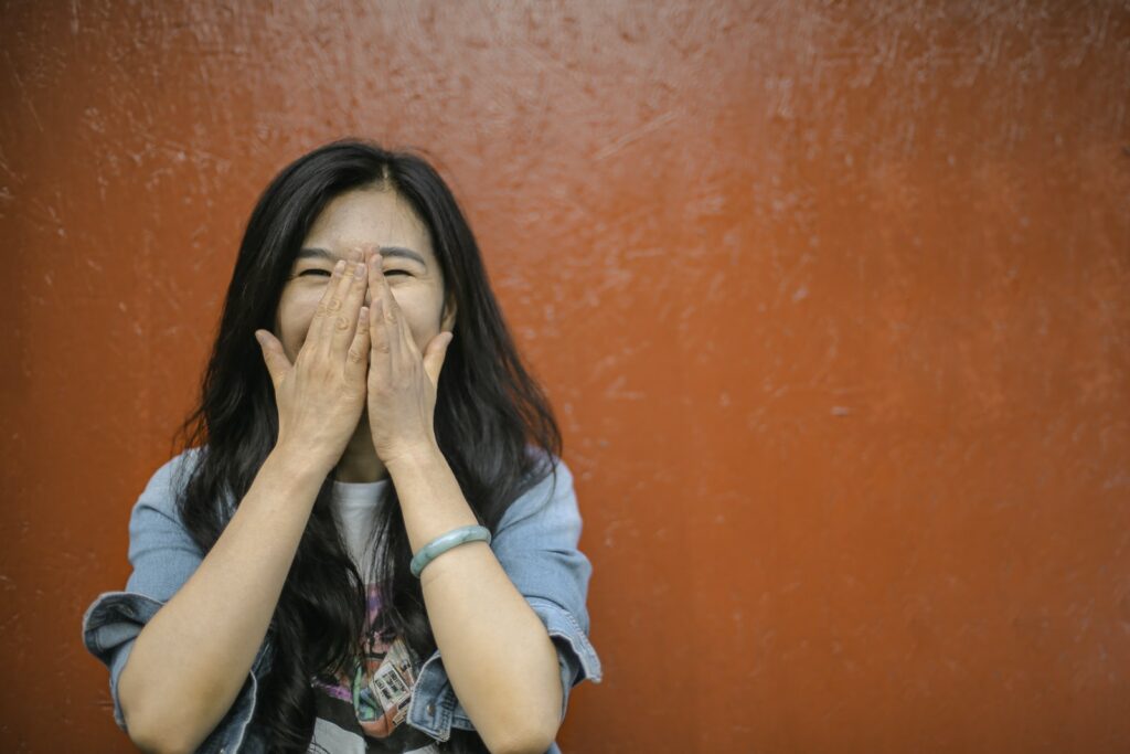 Молодая радостная азиатка в повседневной одежде закрывает лицо руками и смеется, стоя на фоне яркой оранжевой стены