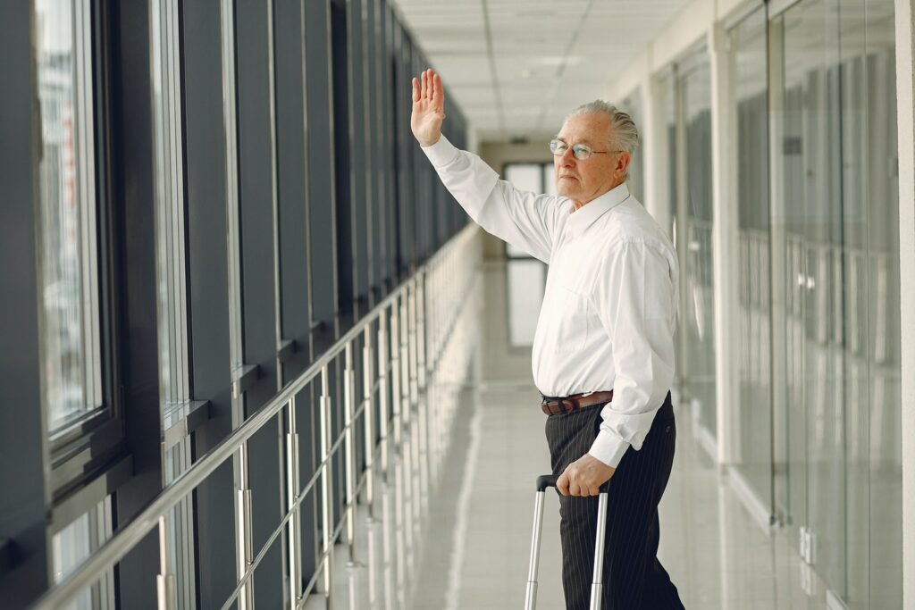Вид сбоку на пожилого спокойного мужчину в официальной одежде с чемоданом, который идет по коридору современного аэропорта и машет рукой на прощание, глядя в стеклянную стену