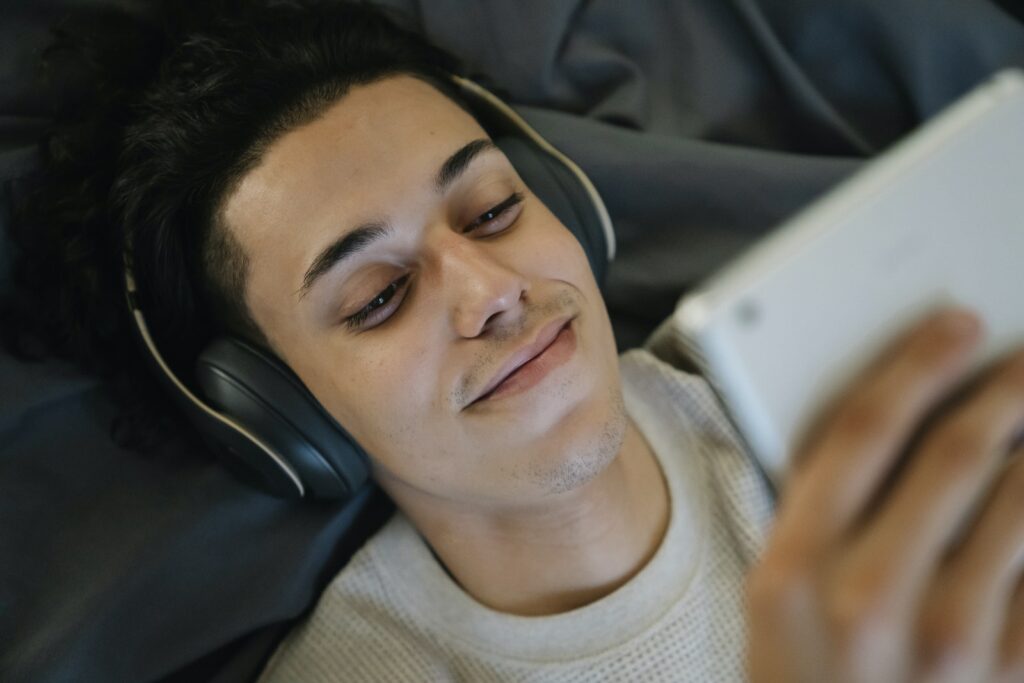 Positiver junger hispanischer Mann mit Kopfhörern, der lächelt und einen Film auf einem Tablet ansieht, während er sich zu Hause auf dem Bett entspannt