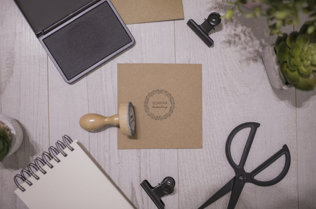 Top view of traditional rubber stamp and ink pad placed on wooden table near spiral notepad and composed with stationery and potted plants