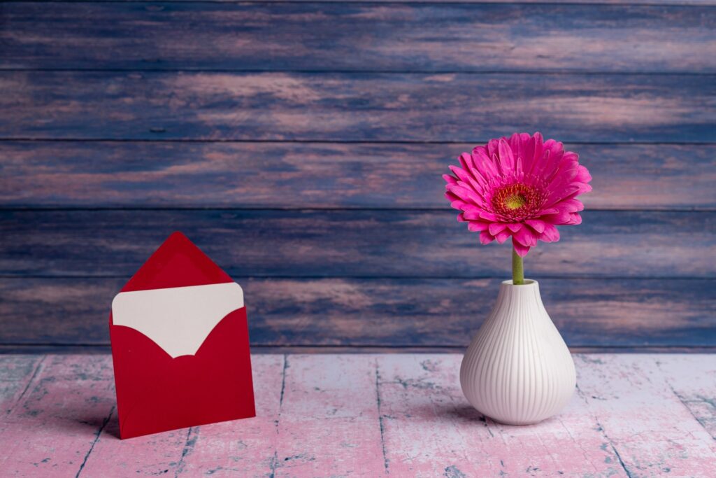 Piccolo vaso con fiore di Gerbera jamesonii rosa disposto con cartolina in busta rossa su tavolo in legno