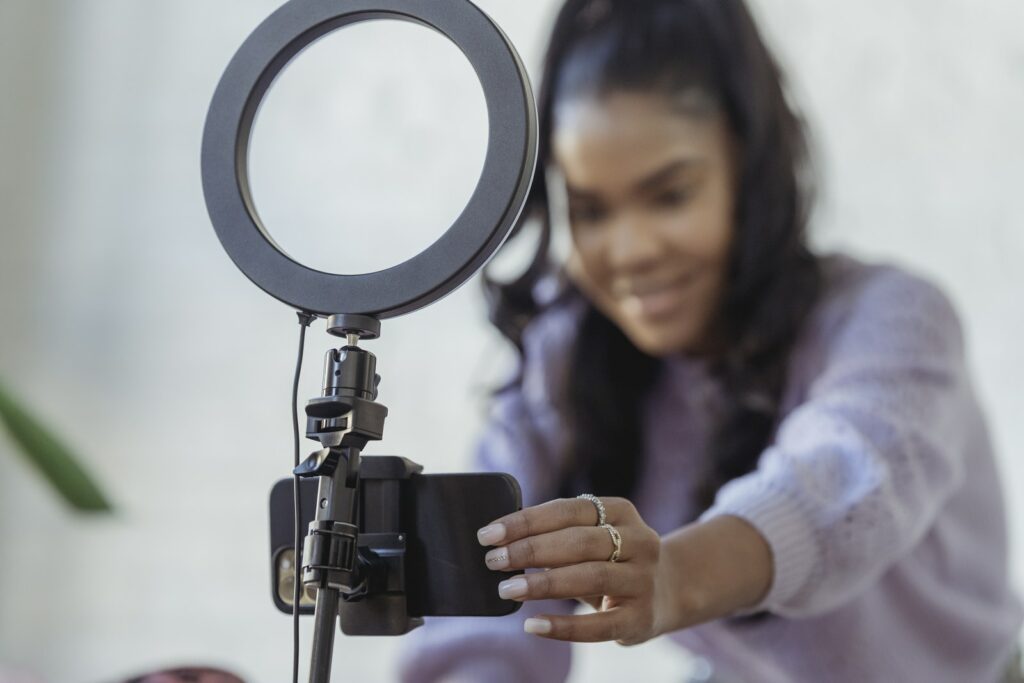 Jovem blogueira afro-americana alegre com uma camisola elegante e sorridente enquanto monta uma câmera de smartphone acoplada ao tripé com luz de anel antes de gravar o vlog