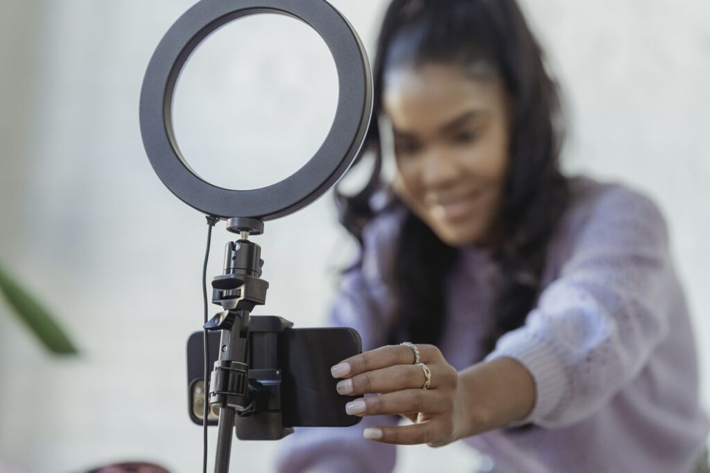 Giovane e allegra blogger afroamericana con maglione alla moda che sorride mentre imposta la fotocamera dello smartphone sul treppiede con luce anulare prima di registrare un vlog