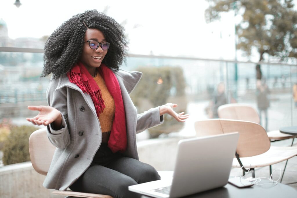 Femme afro-américaine heureuse et excitée, riant et gesticulant lors d'un appel vidéo sur son ordinateur portable, assise dans un café moderne.