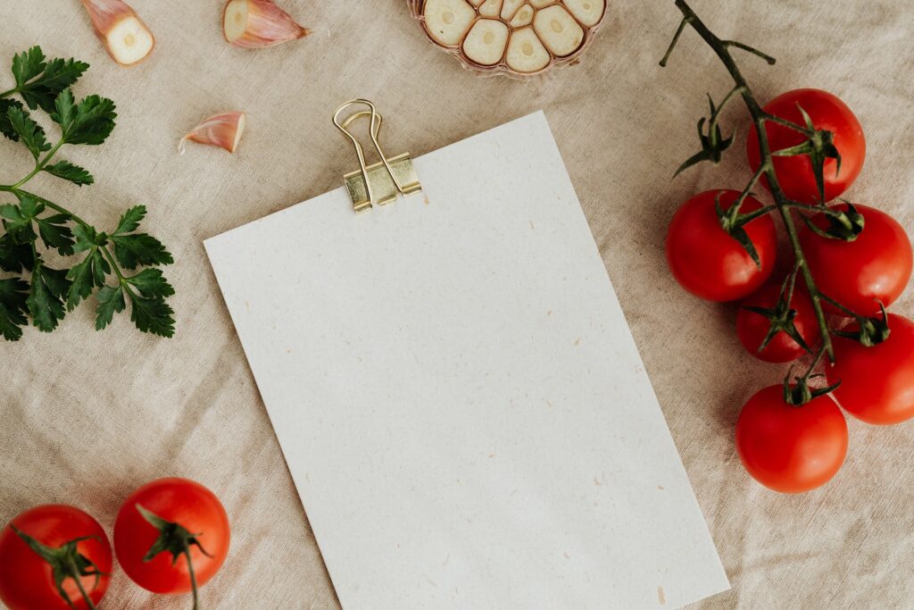 リネンのテーブルクロスの上に置かれた、金色の紙でできたバインダー付きの白紙のクリップボードの上から、枝についた美味しそうな赤いトマトと、レシピやメニューの配置に役立つカットしたニンニクと緑のパセリ。